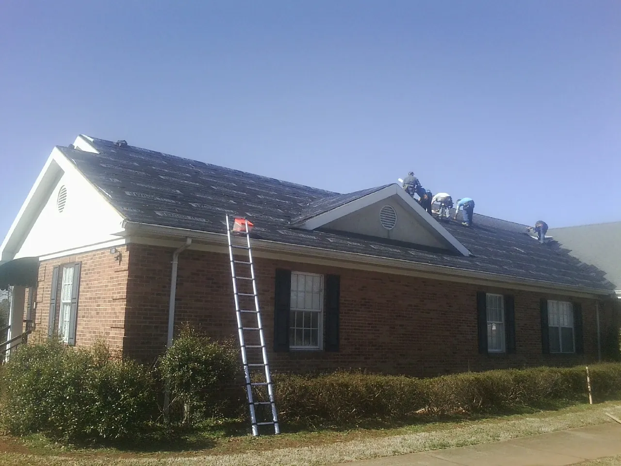 Crew tearing shingles off a brick ranch roof with a ladder set — Metal Roof Repair work by our crew in Hardwick