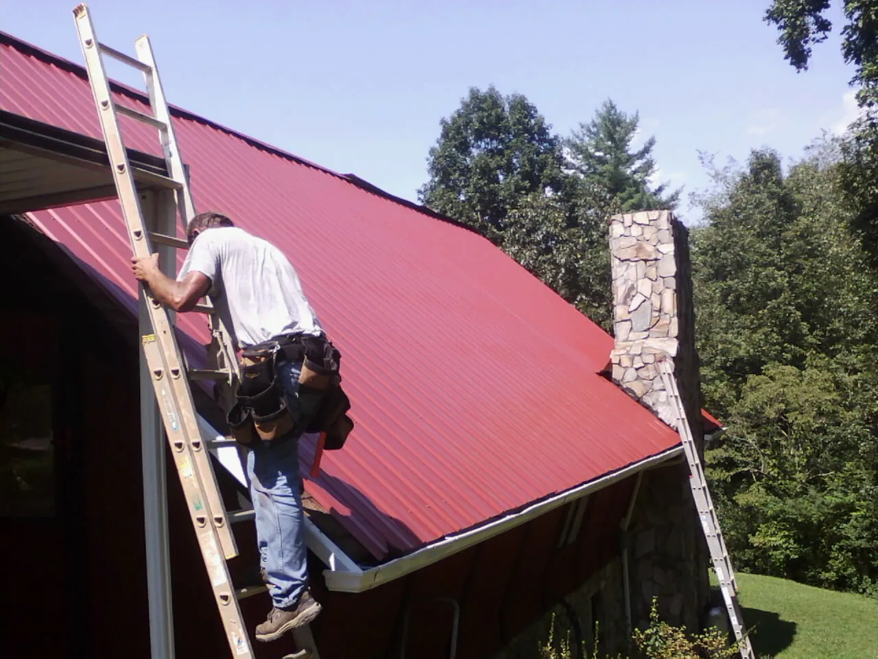 Roofer on a ladder beside a red metal roof and stone chimney — Soffit & Fascia Repair by Roofing Brothers in Hardwick